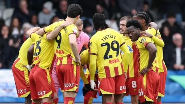 The Watford team gather in a huddle with their arms around each other on the pitch. They wear yellow shirts, red shorts and red socks.