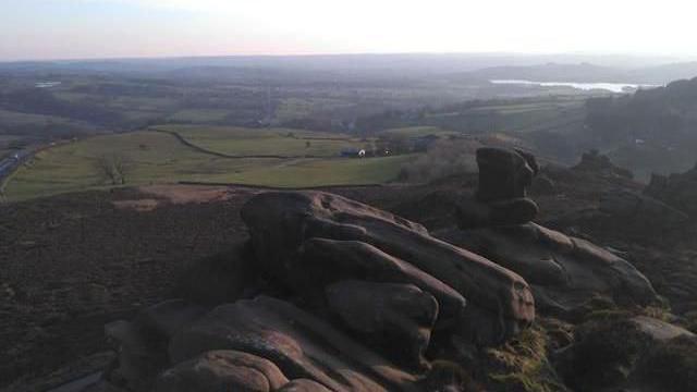 An aerial view of a rocky ridge in the middle of the Peak District. The rocky ridge is in the foreground while a road can be seen snaking through fields in the distance.
