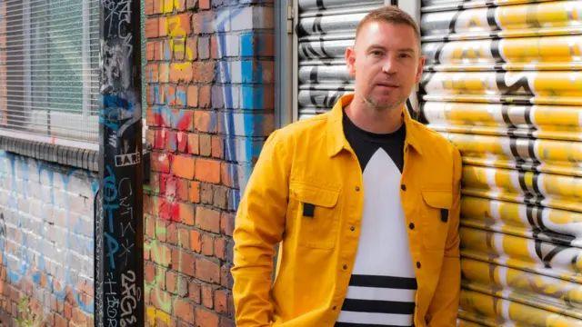 Mark standing outside, wearing a yellow jacket with short hair, standing next to a wall and metal shutters, covered in graffiti on a street 