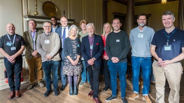 Photo of nine men and three women in a line, all wearing lanyards. They are in a stately home with a stone fireplace and wooden floor.