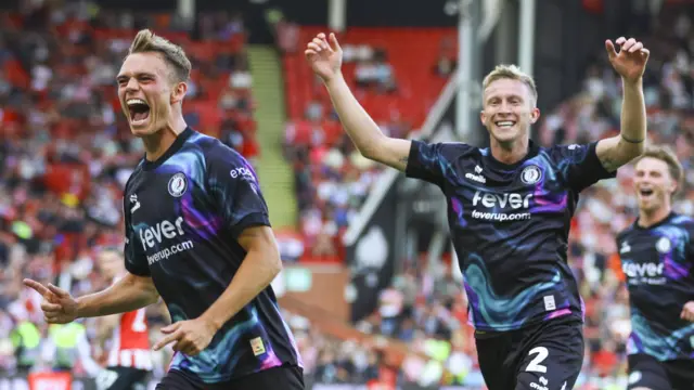 Scott Twine (left) runs with his mouth open smiling while pointing his finger with Ross McCrorie following behind with two arms in the air