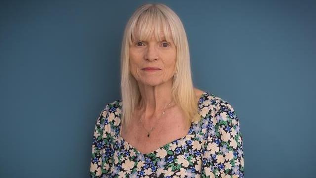 Dr Denise Chaffer - a middle-aged woman with light blonde, shoulder-length hair. She is looking directly at the camera and is wearing a blue, green and cream floral print sleeved dress and is wearing a necklace with a small dark pendant on it. Denise has a neutral expression on her face and is standing in front of an all-blue backdrop