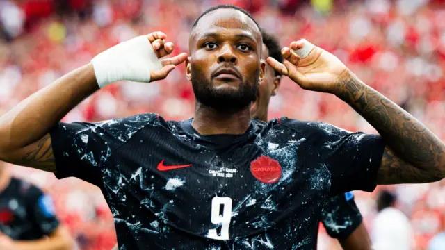 Cyle Larin, wearing Canada's black away strip with white patterns on it, celebrates scoring at the World Cup against Qatar by putting his hands either side of his head and pointing to his ears with his index fingers