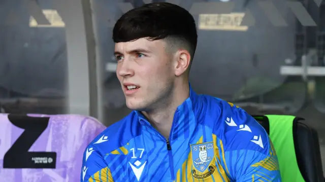 Charlie McNeill sits in the dugout during a game for Sheffield Wednesday. He is wearing a light blue and yellow club tracksuit top.