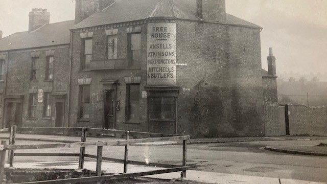 A black and white picture of the Ring O' Bells pub on Yardley Street, on the corner of Nelson Street. It is at the end of a row of buildings and has a sign on which says 'Free House, Ansells, Atkinsons, Worthingtons, Mitchells and Butlers. Opposite the pub is a wooden fence which looks like it is around a brick hole. 