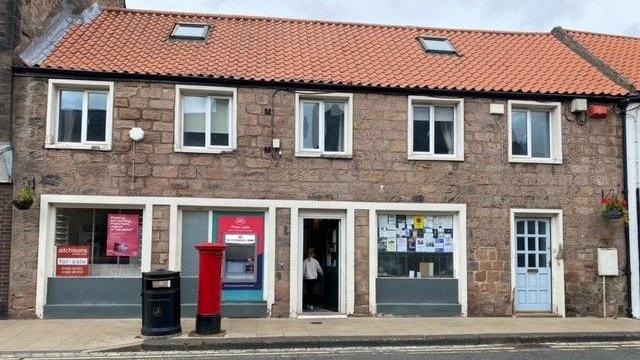 A picture of a post office with a man standing in the doorway and a postbox prominent in the foreground. It's a long two-storey building with three large windows. 