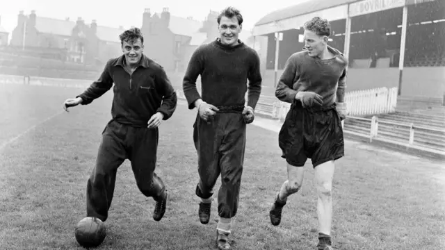 Peter Higham is running with an old leather ball at his feet in a tracksuit, alongside two other players at Nottingham Forest's City Ground in 1956, black and white photo