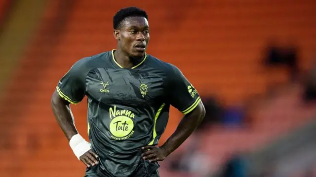Lincoln City striker Tanto Olaofe stands with his hands on his hips during a pause in play. He is wearing a dark grey away shirt.