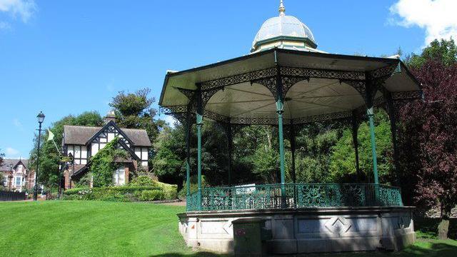 The bandstand photographed in 2014. It has green fence and pillars complete with intricate ornaments. It is surrounded by a freshly-cut grass and trees. There are several multi-storey houses in the distance.