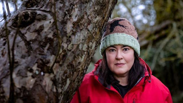 Anna Gazeley is standing by a large trunk of a tree and looking at the camera. She has long dark hair and is wearing a green and brown woolly hat and a red coat