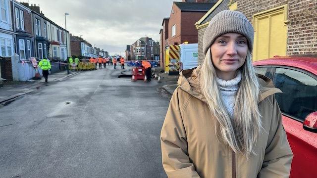 Lucy Mouter stands in the street with the sinkhole behind her. She had long blonde hair and wears a beanie hat.