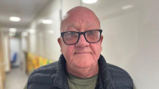 A 67 year old bald man with large, dark framed glasses, looking directly at the camera, weraing a navy blue puffer gilet and a green top, standing in the corridor of the scanning lorry.