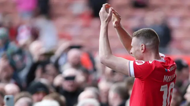 Wearing a red Wrexham shirt Paul Mullin, facing away from camera, applauds Wrexham fans on the pitch