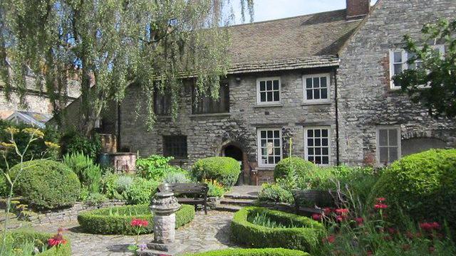 An ornate garden behind Scaplen's Court, a two-storey historical building. The garden has sculptured hedges, paving and trees,