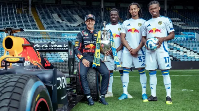 Willy Gnonto, Largie Ramazani and Isaac Schmidt of Leeds United alongside the Championship trophy and Red Bull driver Arvid Lindblad.