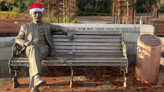 A bronze statue of John J.R. Macleod sitting on a bench with a red and white santa hat on its head. The bench is brown and wood coloured and has been weathered slightly. The statue is in a seated position with its left hand resting on the top of the bench. A marble wall is behind with an inscription reading "...All owe their lives to Insulin".