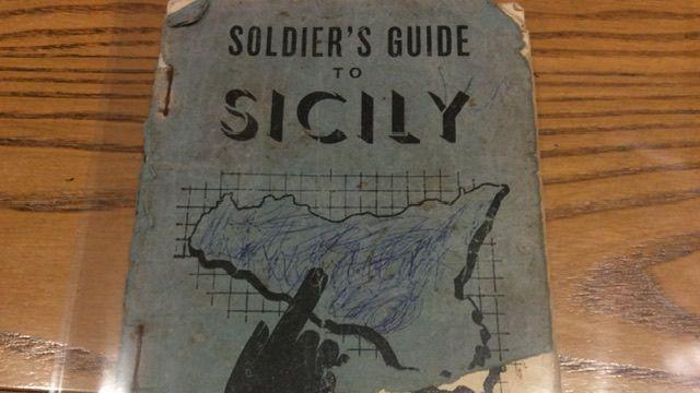 The Soldier's Guide to Sicily Handbook. It's a muted blue colour with worn edges. The cover text is black and there is an outline of Sicily with a black printed finger pointing to the centre of it.