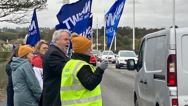 Several protesters at the roadside carrying blue banners with the NASUWT logo. A van is passing them on the right-hand side.