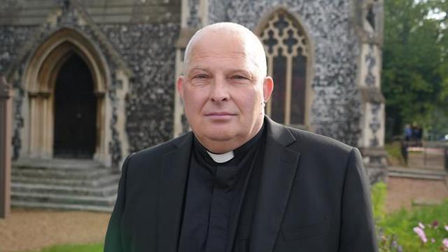 A man standing in front of the church looking at the camera. He is wearing black 