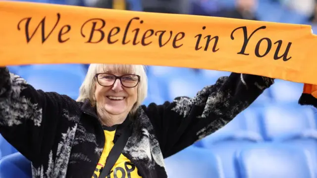 A Wolves fan holds a scarf saying "We believe in you"