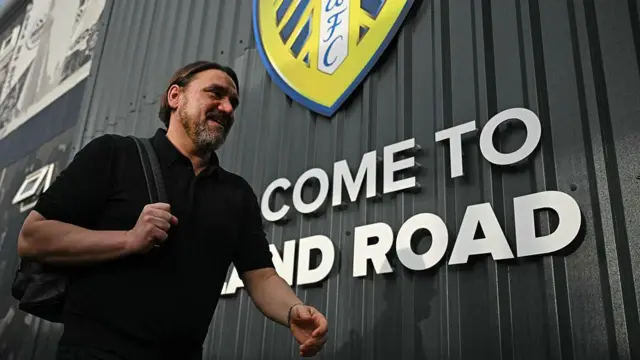 A smiling Dabiel Farke with a rucksack over his shoulder, walking past a "welcome to Elland Road" sign