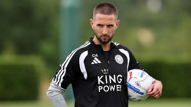 Conor Hourihane wearing a black Leicester City training top with King Power written across the middle and holding a football under his arm while watching training