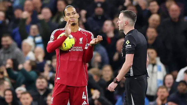 Virgil van Dijk, holding the ball during Liverpool's loss to Manchester City and sarcastically applauding in the direction of referee Chris Kavanagh. The corwd is out of focus in the background.