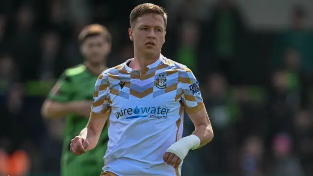 Matt Curley is wearing a white Tamworth FC away shirt and is about to pass the ball. The ball is out of shot. Curley is also wearing a white bandage on his left wrist. An opposition player is in green in the background but the image is blurred.