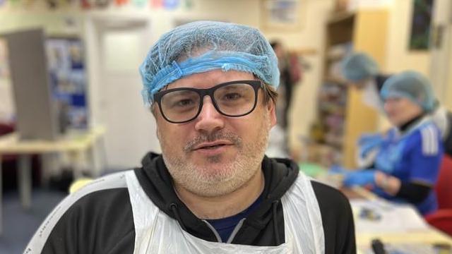 A man smiles at the camera and has a blue hair net over his head. He has black glasses and is wearing a black hoodie. He has a plastic vest like cover over his torso to protect his hoodie.