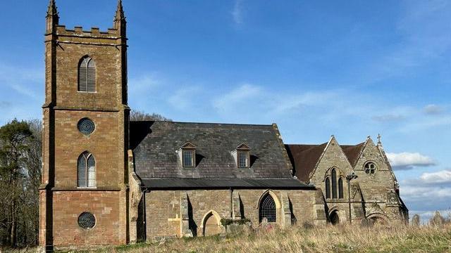 A two-storey church with three story tower and a pitched slate roof with dormer windows. It is in a field, with tall grass. The picture is taken on a sunny day, with light clouds and blue sky.
