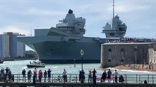 The large carrier close to the shore next to a tug boat with people gathered to watch it on its journey