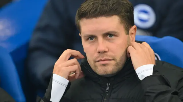 Brighton manager Fabian Hurzeler adjusts his collar while sitting in the dugout