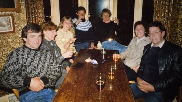 A group of three men and three women sat around an oblong pub table with drinks in the 1980s