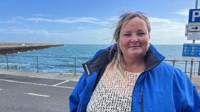 Donna De Gruchy stood in front on the sea front. She is wearing a white knitted top with a blue coat. She is looking at the camera with her hair blowing in the wind. She has sun glasses on her head. Behind her is the sea and a grey road above with railings. 