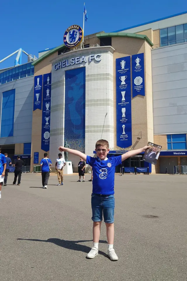 Young boy standing with arms aloft outside of Stamford Bridge