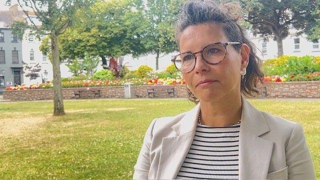 Susana Rowles in Parade Gardens wearing a smart jacket with striped top under and glasses looking to the left of the frame, in the background the gardens, flower beds and trees are visible 