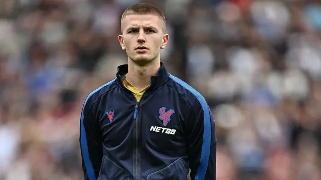 Adam Wharton, wearing a Crystal Palace training top, lining up before a match, with the crowd a blur behind him