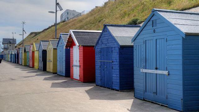 A row of blue, red, yellow and green beach huts. There is a path in front and a steep grassy slope behind.