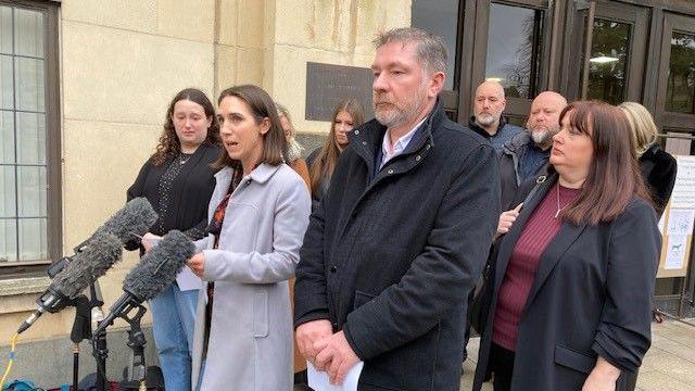 A group of people stood in front of microphones. Some are holding sheets of paper. They are in front of Oxford Crown Court