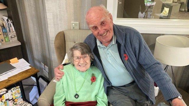 Michael Woods with his wife Mary. They are seated in an armchair with Michael perched on the arm. he gives his wife a hug. Both are smiling and wearing Remembrance poppies on their jumpers.