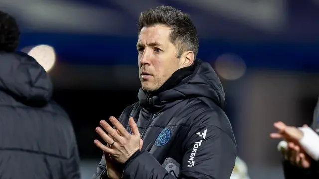 QPR boss Julien Stephan, wearing a navy blue coat with the QPR badge on it, is clapping.