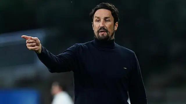 A bearded Cesar Peixoto pointing at something on the pitch with his right index fingetr while wearing a dark blue roll neck pullover