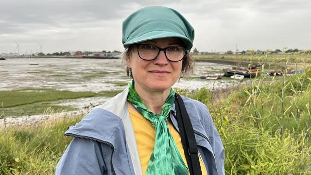 A woman in a green hat and green scarf. She is also wearing a yellow shirt and grey coat. She is standing in what looks like a marsh.