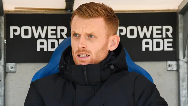 A close-up of Edward Still, wearing a black zip-up jacket, sitting on a blue chair in the dugout in Genk
