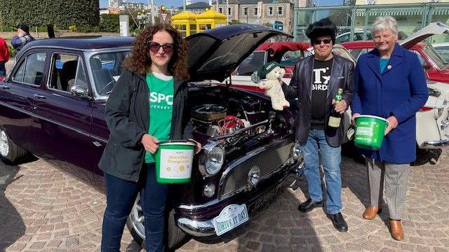Two women and a man stand beside a classic car with its bonnet open. The two women are holding green fundraising buckets. The man is holding a bottle of unopened Champagne and a teddy bear in a green hat.