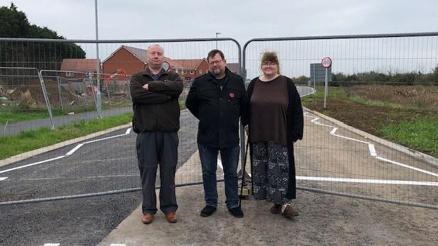 three people standing in front of fencing with new road and housing development in the background.