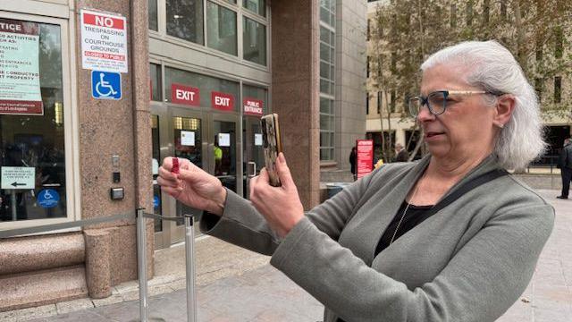 Peggy Savani holds up a red raffle ticket and takes a photo of it with her cell phone outside the a marble courthouse. Trees are seen in the distance and Peggy is wearing glasses and a grey cardigan. 