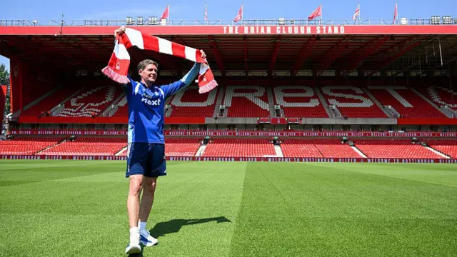 Oliver Glasner holding a Nottingham Forest scarf above his head on the pitch at an empty City Ground. The Brian Clough Stand is behind him.