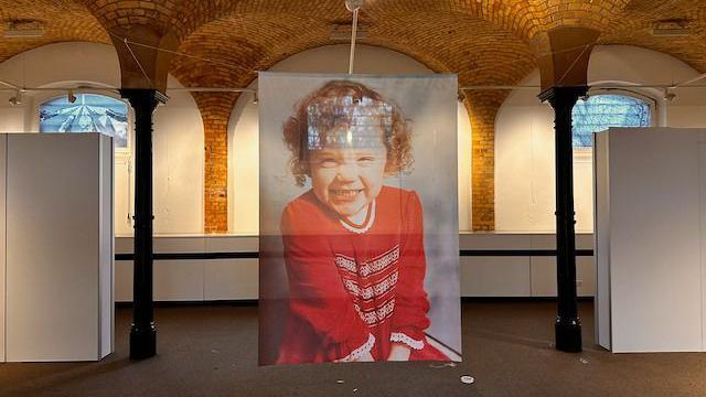 A large semi-transparent photo of Katrice in between two black columns at the art gallery. She is smiling at the camera and has short curly hair. She is wearing a long-sleeved red dress with white collar.