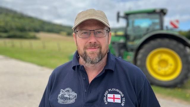 Will Tupper in an England ploughing team t shirt. He stands in front of a tractor.